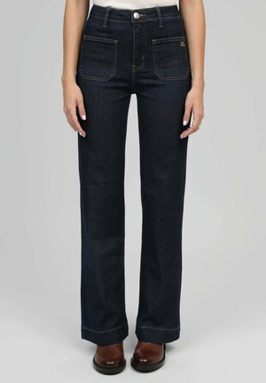 Personne portant un jean droit bleu foncé avec des poches plaquées à l'avant et des bottes en cuir marron, debout devant un fond uni.