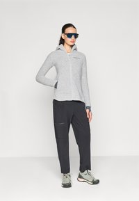 Haut de survêtement en polaire zippé gris avec un design ajusté, associé à un pantalon cargo noir doté de poches zippées et d'une coupe décontractée. Les chaussures sont gris et noir.
