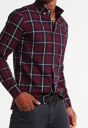 Homme portant une chemise à carreaux rouge et bleu marine boutonnée, rentrée dans un jean sombre avec une ceinture noire, main dans la poche.