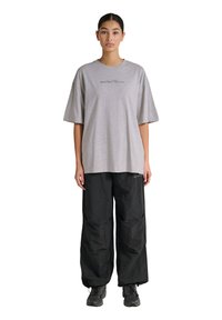 Kvinde står foran, iført en løs grå T-shirt med tekst og løstsiddende sorte bukser samt sorte sneakers på en hvid baggrund.