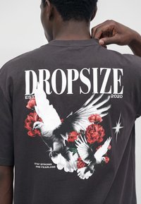 Homme portant un t-shirt foncé avec un texte blanc « DROPSIZE » et « Stay Strong and Fearless », avec un graphisme de colombes blanches et de roses rouges dans le dos.