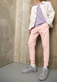 Růžové tepláky Nike, šedé tenisky, levandulová košile, bílá košile na knoflíky. Hladké textury a neformální design, umístěné na neutrálním pozadí.