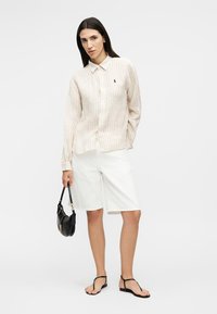 Femme portant une chemise à boutons beige rayée et un short blanc jusqu'aux genoux, tenant un sac à main noir et portant des sandales noires.