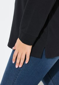 Camisa negra de manga larga con puños acanalados, aberturas en los costados y un corte holgado, combinada con jeans de color azul oscuro. Textura de algodón suave.