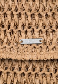 Borsa in paglia intrecciata di colore marrone chiaro con una superficie leggermente texture. Presenta un'etichetta in metallo argentato incisa con "ROXY."