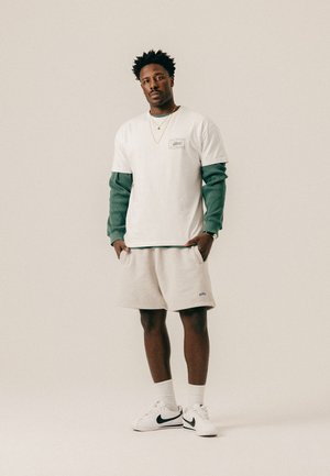 Hombre con cabello corto y rizado que lleva una camiseta blanca de capas, una camiseta de manga larga verde debajo, pantalones cortos beige, calcetines blancos y zapatillas blancas con el logo negro.