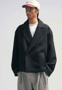 Black wool-blend jacket with a notched lapel, single button closure, and front pockets, paired with a white t-shirt and gray trousers.