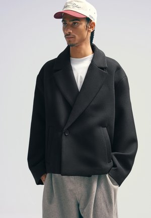 Giacca nera in misto lana con rever a V, chiusura a bottone singolo e tasche frontali, abbinata a una t-shirt bianca e pantaloni grigi.