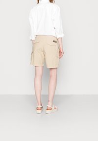 Persona che indossa pantaloni cargo beige, una camicia bianca con maniche arrotolate e sandali con piattaforma beige, in piedi su uno sfondo uniforme.