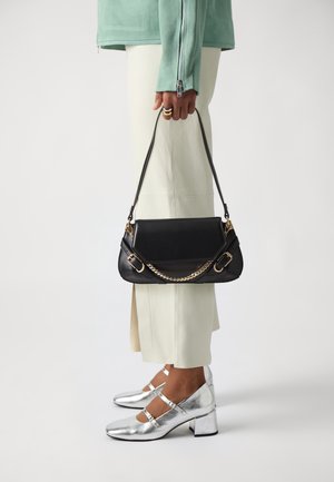 Persoon met witte broek en zilveren hakken houdt zwarte handtas met gouden kettinkdetails naast zich vast tegen een effen achtergrond.