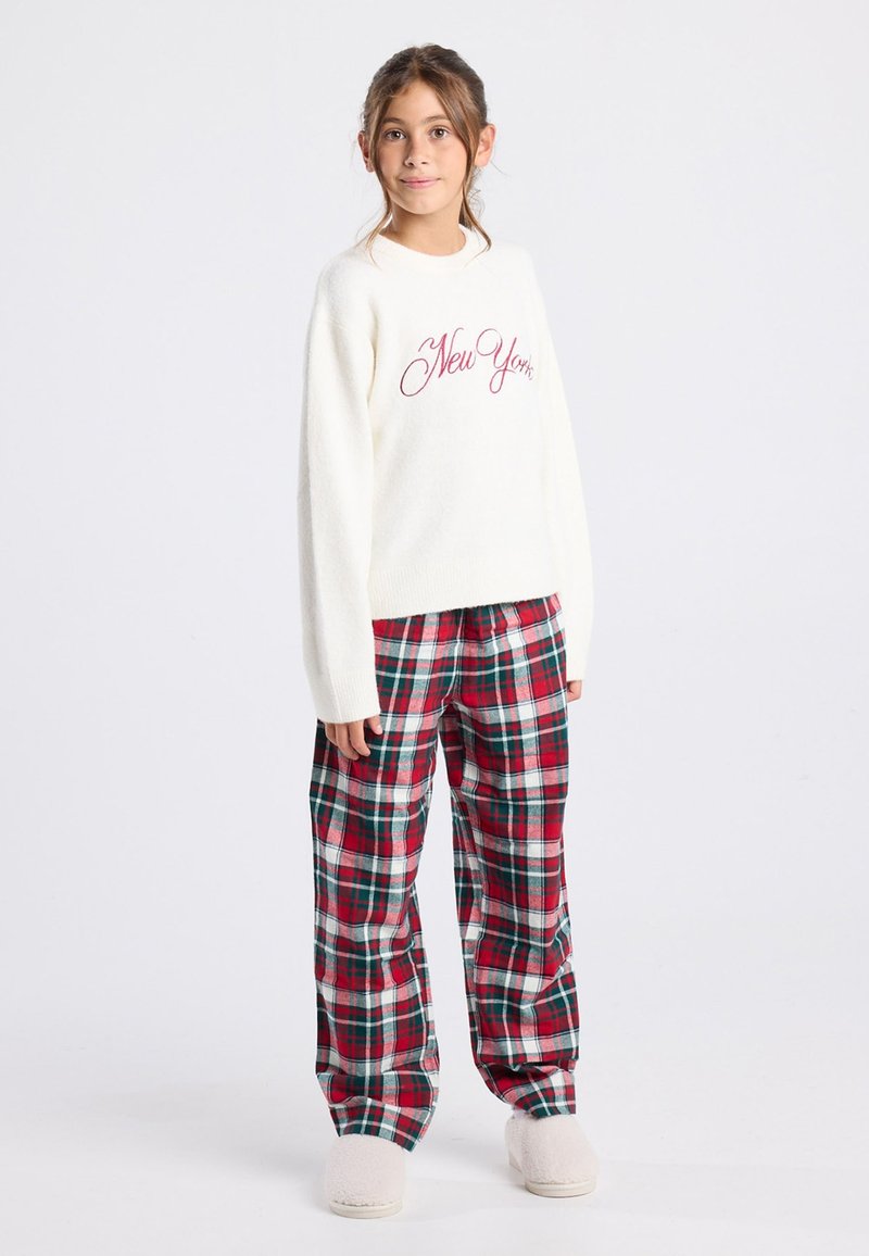 Pull blanc en tricot avec "New York" en cursive rouge. Pantalons de pyjama à carreaux rouges et verts, amples. Chaussures slip-on beiges douces.