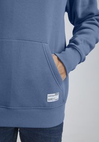 Felpa blu con tasca a marsupio, orlo e polsini a coste, caratterizzata da un'etichetta tessuta sul davanti in basso, realizzata in morbido materiale misto cotone.