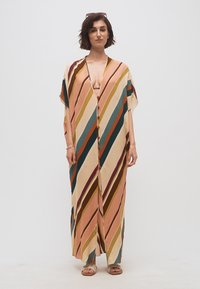 Γυναίκα που φοράει ένα μακρύ φόρεμα με πολύχρωμες διαγώνιες ρίγες και κοντά μανίκια, στέκεται μπροστά σε απλό λευκό φόντο.