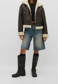 Veste en cuir marron courte avec un col en shearling crème, associée à un short en denim bleu clair. Bottes noires montantes avec des accents en métal argenté.