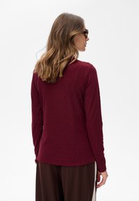 Kobieta z włosami do ramion w jasnym odcieniu brązu, nosząca okulary przeciwsłoneczne, teksturowaną bordową bluzkę z długim rękawem i ciemne szerokie spodnie, odwrócona plecami.