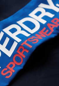 Tissu de sport bleu avec un grand texte blanc et rouge, "SUPERDRY", et "SPORTSWEAR". Texture lisse, vue en angle révélant un matériau superposé.