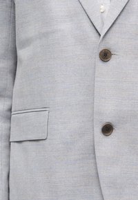 Ανοιχτό γκρι σακάκι κοστουμιού με σχέδιο ψαροκόκαλο, με δύο σκούρα κουμπιά και τσέπη με καπάκι στην αριστερή πλευρά, φορεμένο πάνω από ένα λευκό πουκάμισο με κουμπιά.