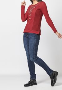 Haut rouge à manches longues texturé avec détail brodé, associé à un jean skinny bleu foncé et des bottines noires à lacets.