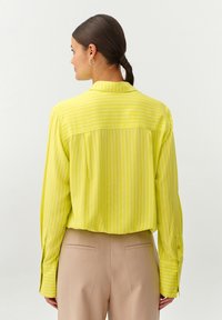 Camisa abotonada a rayas amarillas con mangas largas, corte relajado y detalle en el yugo trasero. Combinada con pantalones beige ajustados.