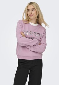 Roze sweatshirt met de tekst "Harvest" in een glanzend, kleurrijk patroon. Zachte stof, ronde halslijn, geribbelde manchetten en zoom. Gedragen over een witte top.
