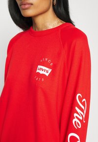 Czerwona bluza z miękkiego materiału, z białym logo Levi's na klatce piersiowej i napisem "The" w kursywie na prawym rękawie.