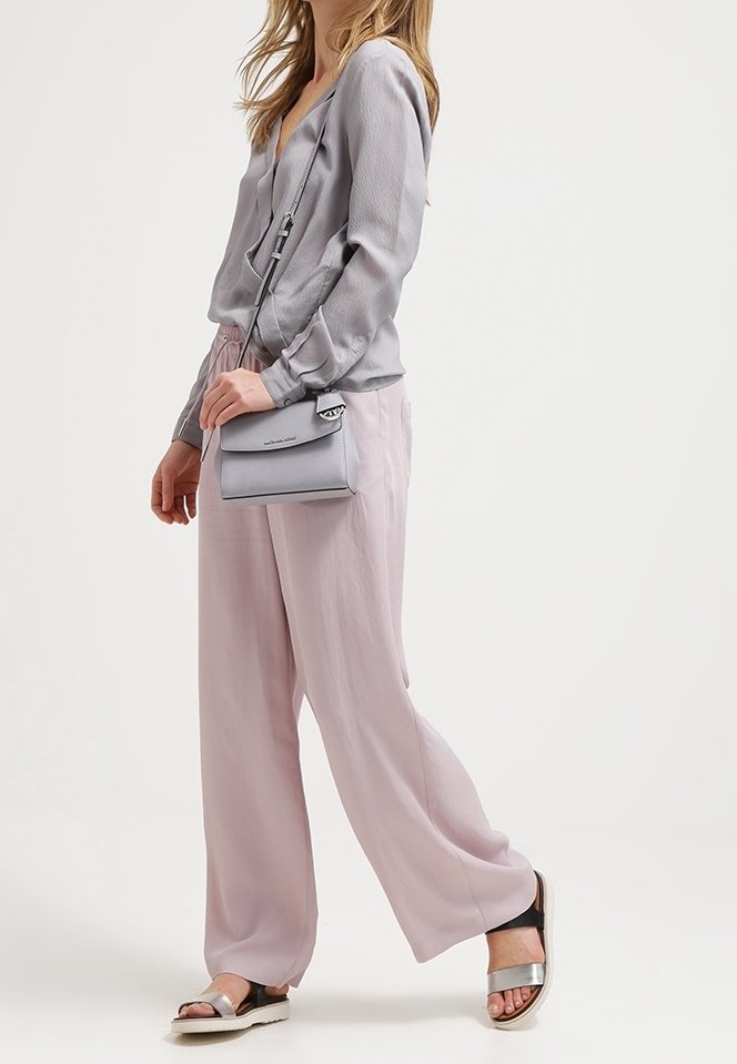 Femme portant un chemisier gris clair et un pantalon large rose, portant un petit sac gris en bandoulière et des sandales argentées.