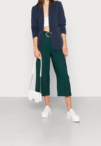 Blazer bleu marine, crop top blanc, pantalon vert à jambes larges, baskets à plateforme blanches, et sac à main blanc avec bandoulière chaîne.