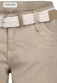 Beige Baumwollhosen mit geschwungenen Fronttaschen, kombiniert mit einem hellen gewebten Gürtel mit einer silbernen Metallschnalle und einem braunen Lederakzent.