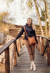 Schwarze Puffermantel, braune Leder-Schultertasche, beige Leggings und beigefarbene Stiefeletten. Holzbrücke mit natürlichen Hintergrundelementen.