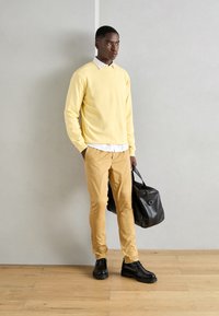 Gelber Pullover, weißes Hemd mit Kragen, beiger Hose und schwarze Schuhe. Schwarze Ledertasche mit einem runden Griff, vor einem neutralen Hintergrund.