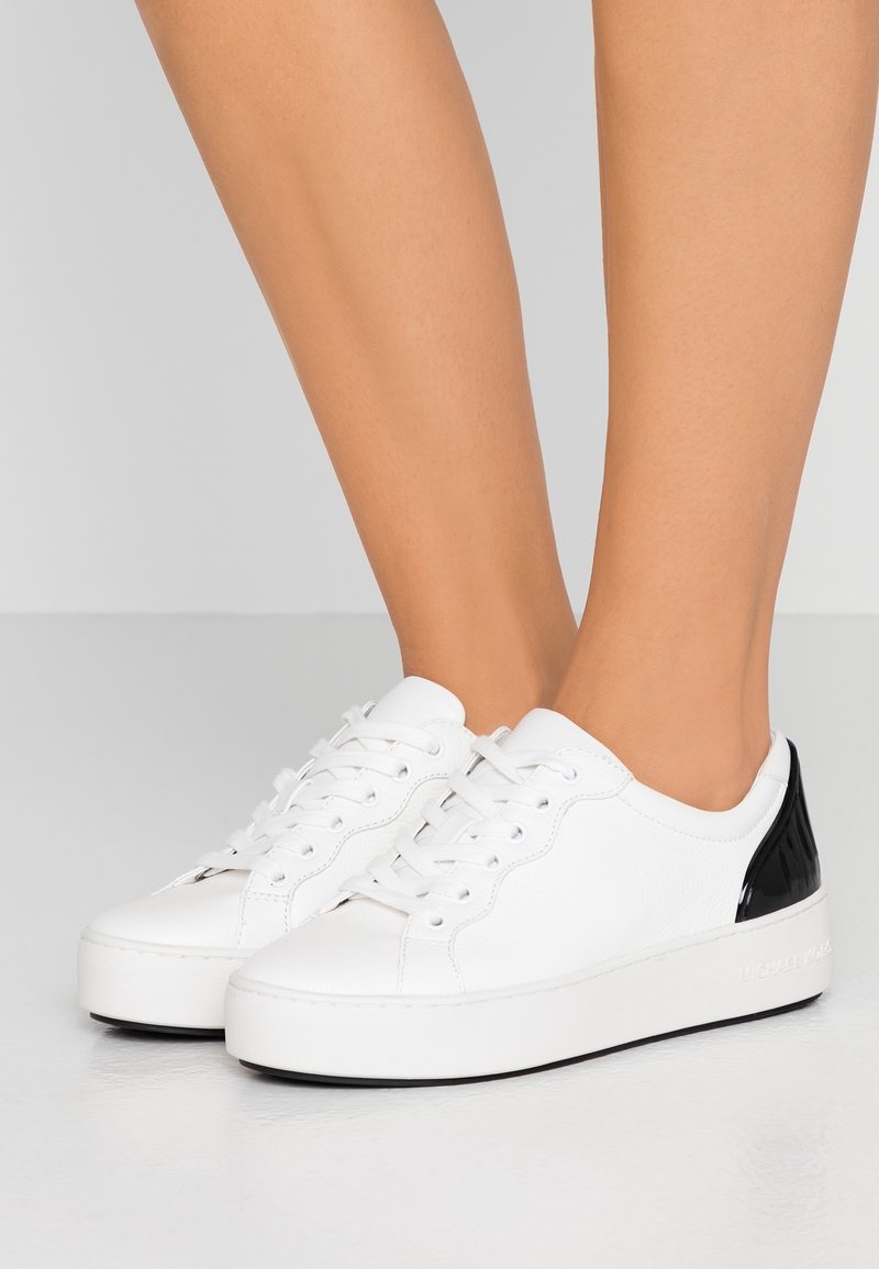 Baskets en cuir blanc avec un accent verni noir au talon, dotées d'un bout arrondi, d'un laçage à l'avant et d'une surface texturée.