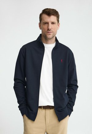 Man met kort bruin haar, gekleed in een marineblauwe jas met rits, wit T-shirt en beige broek, staand met handen in de zakken.