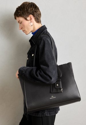 Black leather tote bag with textured finish and a detachable strap, displayed alongside a person wearing a black jacket and blue top.