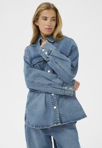 Chemise en denim bleu clair, dotée de grandes poches avant, avec des accents de boutons en métal argenté, et une coupe décontractée avec des épaules tombantes et un ourlet arrondi.