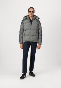 Grå pufferjakke med lynlås foran og hætte, med en quiltet design og kontrasterende mørkegrå skulderpaneler. Båret med mørke jeans og sorte sneakers.