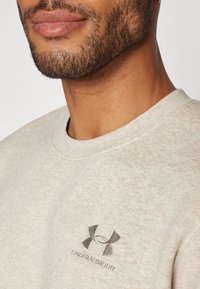 Närbild av en mans underansikte och nacke som bär en ljusbeige Under Armour-tröja med en synlig logotyp på bröstet.