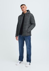 Grå quiltet jakke med hætte, med lynlås og sidelommer, båret med mørke jeans og hvide sneakers med sorte striber.