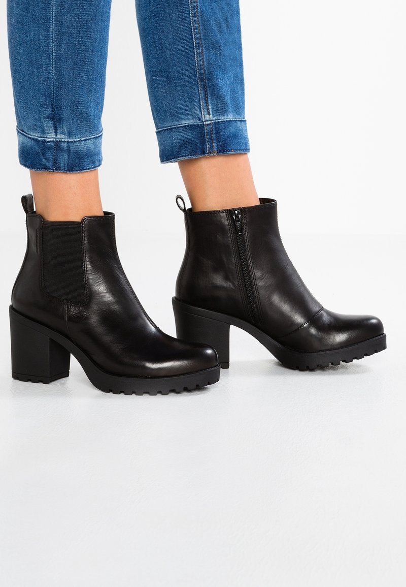 Bottines en cuir noir à talons blocs avec fermeture éclair latérale portées avec un jean bleu retroussé sur fond blanc.