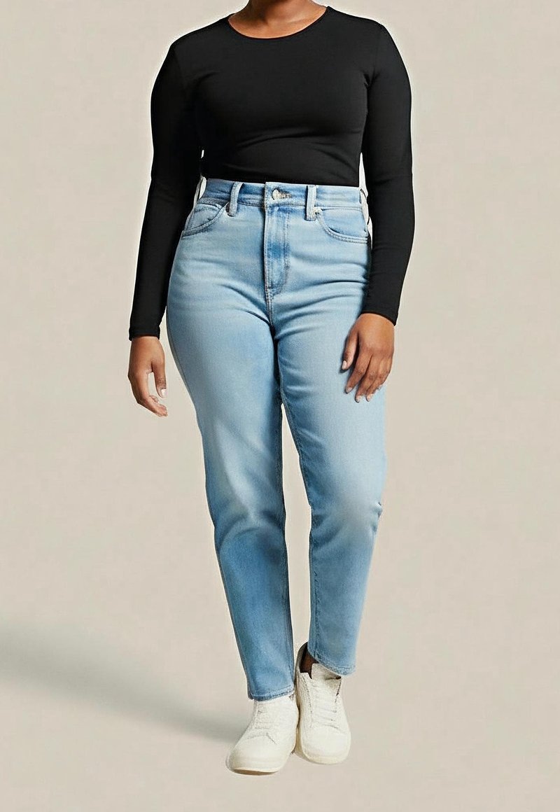 Personne portant un jean taille haute bleu clair, un haut à manches longues noir et des baskets blanches, debout devant un fond uni.