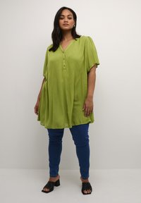 Tunica verde lime a maniche corte con scollo a V e tre bottoni, abbinata a jeans skinny blu e sandali neri con tacco.