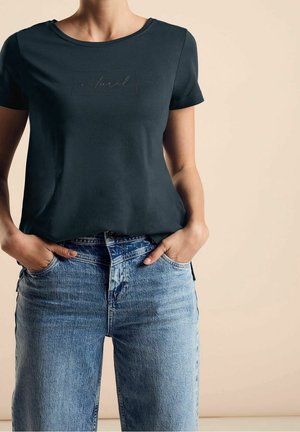 Femme portant un t-shirt noir ajusté avec le texte « natural » et un jean bleu taille haute, mains dans les poches avant, fond beige.