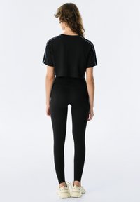 Zwarte cropped t-shirt met blauwe zijstrepen, gecombineerd met zwarte high-waisted leggings. Model draagt lichtgekleurde sneakers.