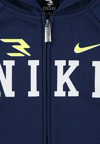 Felpa con zip di colore navy con grande scritta bianca "NIKE", un logo Nike giallo e hardware della zip argentato, dotata di cappuccio. Materiale in cotone morbido.