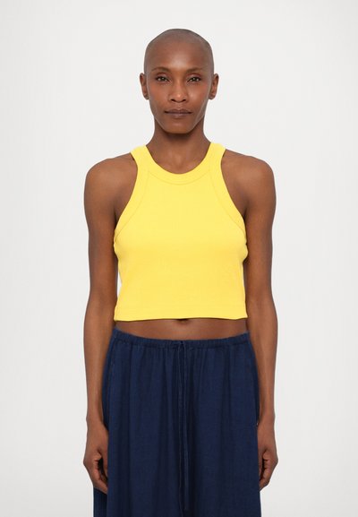 Gelbes geripptes Crop-Top mit hohem Kragen und breiten Trägern, kombiniert mit lockeren, navyblauen Hosen. Weiche Textur und taillierter Schnitt.