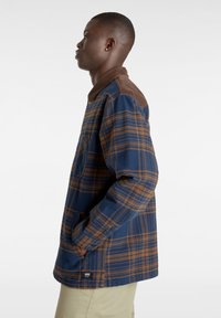 Rutigt skjorta i blått och brunt, tillverkad av tjockt tyg. Har en krage i brunt kordflöjt, långa ärmar och en sidoficka.