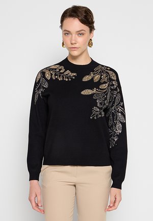 Mujer con suéter negro con bordado floral en oro y plata en los hombros y mangas, combinado con pantalones beige y pendientes de aro dorados.