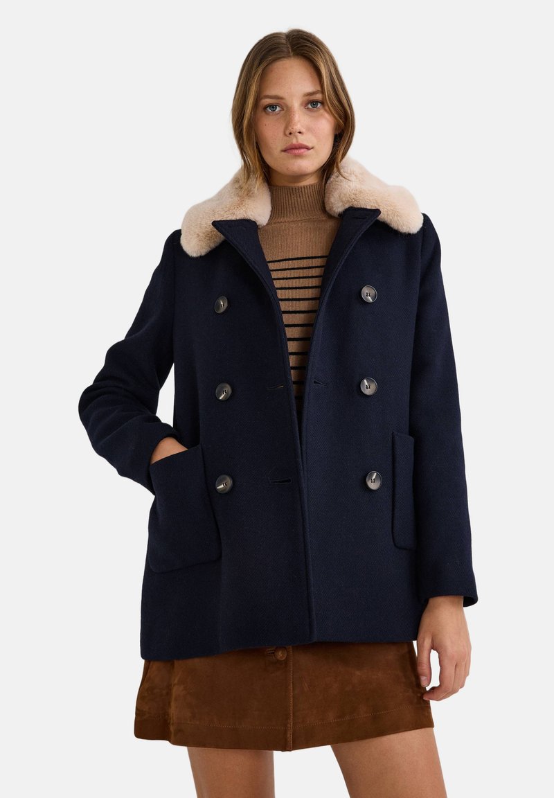Manteau en laine marine avec un col en fausse fourrure pâle, design à double boutonnage, poches avant et boutons. Le modèle porte un pull rayé marron en dessous.