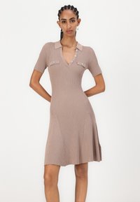 Ribbet, beige kjole med V-hals, korte ermer og dekorative knapper. Har to frontlommer og utvidet hem. Glatt tekstur.