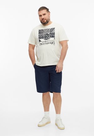 Mężczyzna z brodą, ubrany w biały t-shirt z grafiką, granatowe szorty, białe skarpetki i białe sneakersy, stojący z rękami częściowo w kieszeniach.