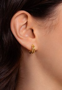 Orecchino a cerchio in oro con un design testurizzato a onde. Presenta una superficie liscia e una finitura lucida, indossato sul lobo di un orecchio.